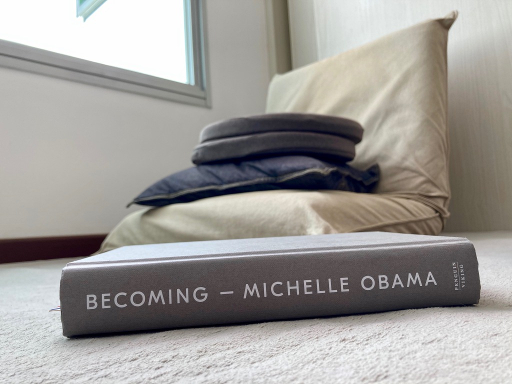 Book lying on floor. The book spine has the words "Becoming - Michelle Obama". In the background is a floor chair and part of a window. 